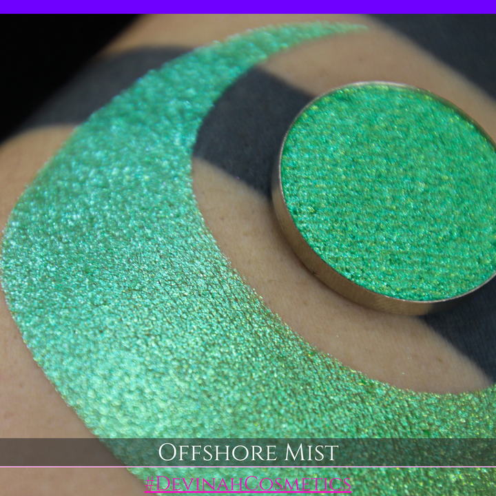A round eyeshadow pan labeled 'OFFSHORE MIST' made by Devinah Cosmetics, placed over a large crescent moon swatch of the same shimmering shifty eyeshadow on skin. The eyeshadow has a duochrome multichrome shift between colors lime, green, yellow, blue. 