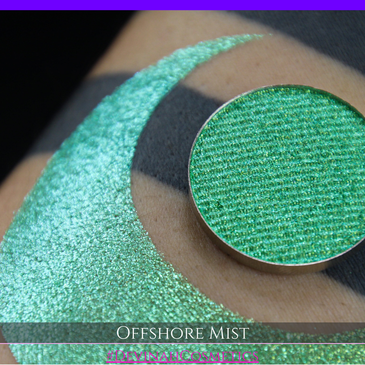 A round eyeshadow pan labeled 'OFFSHORE MIST' made by Devinah Cosmetics, placed over a large crescent moon swatch of the same shimmering shifty eyeshadow on skin. The eyeshadow has a duochrome multichrome shift between colors lime, green, yellow, blue. 