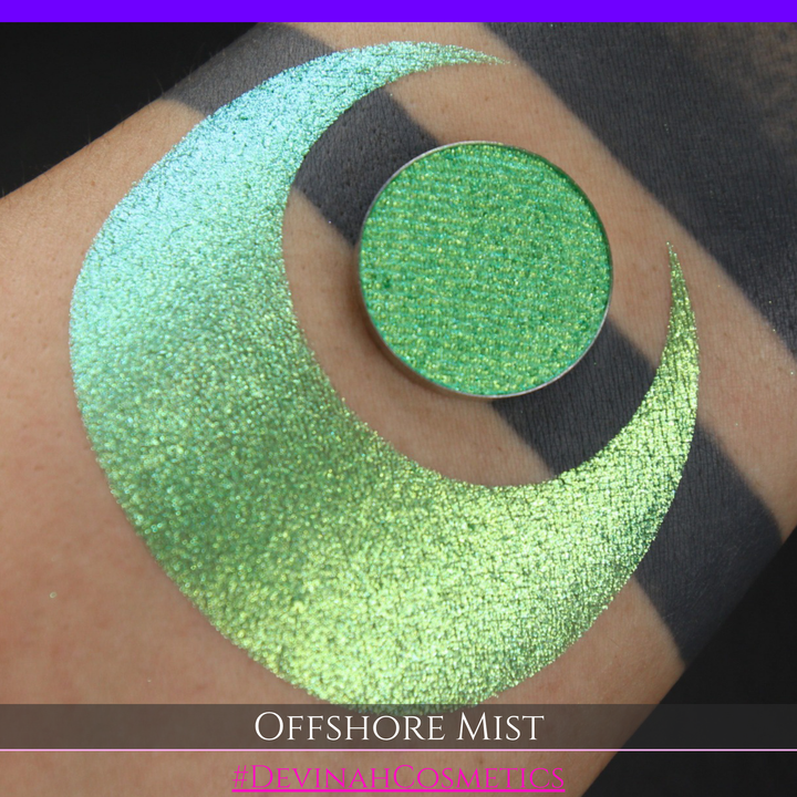 A round eyeshadow pan labeled 'OFFSHORE MIST' made by Devinah Cosmetics, placed over a large crescent moon swatch of the same shimmering shifty eyeshadow on skin. The eyeshadow has a duochrome multichrome shift between colors lime, green, yellow, blue. 