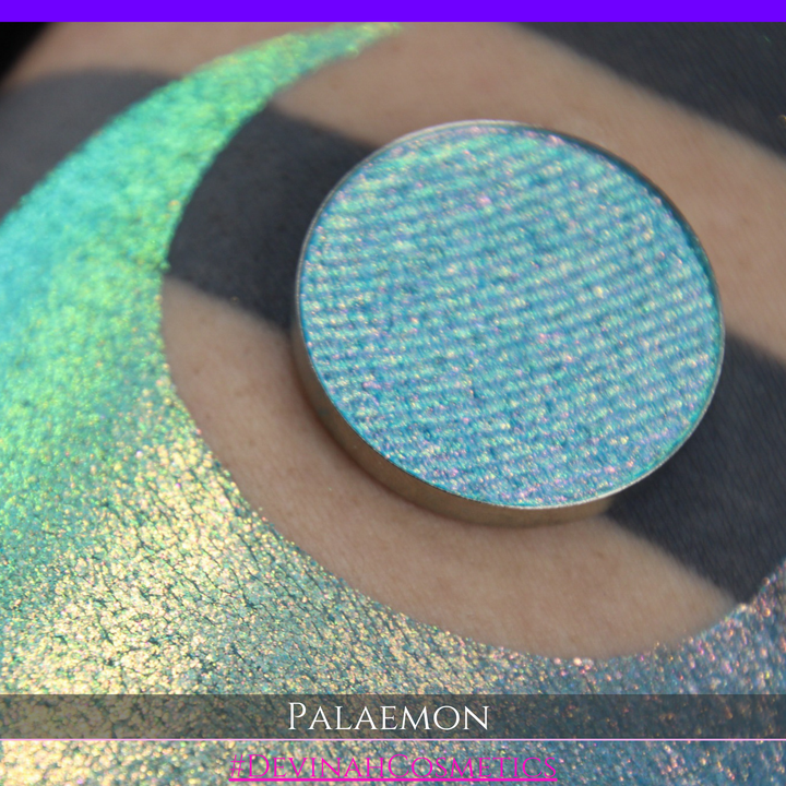 A round eyeshadow pan labeled 'PALAEMON' made by Devinah Cosmetics, placed over a large crescent moon swatch of the same shimmering shifty eyeshadow on skin. The eyeshadow has a duochrome multichrome shift between colors lime green, teal, yellow, gold, orange, pink, red.