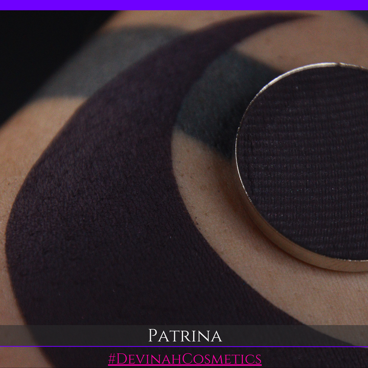A close-up of a makeup eyeshadow pressed powder pan showing a dark charcoal/black plum pigment, with a swatch of the same product on skin. The text 'PATRINA' and '#DEVINAHCOSMETICS' are displayed at the bottom.