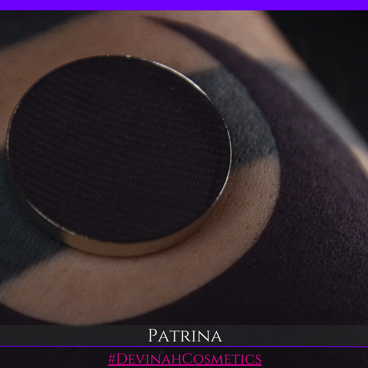 A close-up of a makeup eyeshadow pressed powder pan showing a dark charcoal/black plum pigment, with a swatch of the same product on skin. The text 'PATRINA' and '#DEVINAHCOSMETICS' are displayed at the bottom.