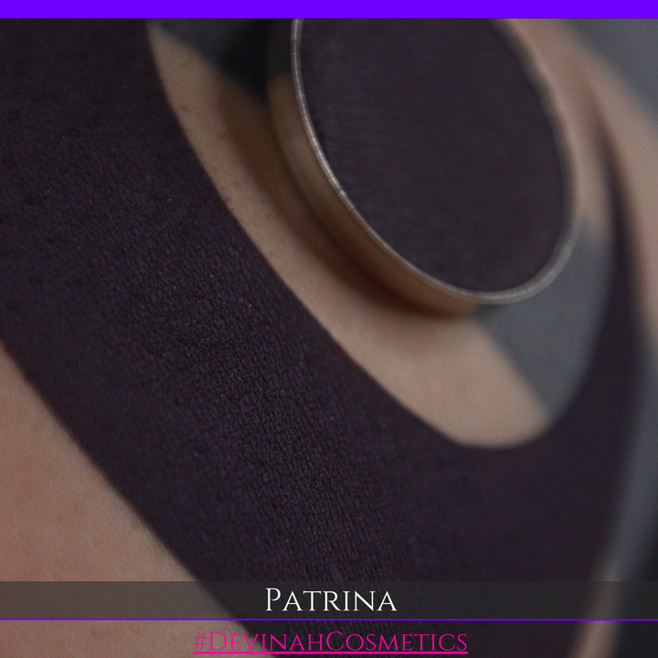 A close-up of a makeup eyeshadow pressed powder pan showing a dark charcoal/black plum pigment, with a swatch of the same product on skin. The text 'PATRINA' and '#DEVINAHCOSMETICS' are displayed at the bottom.