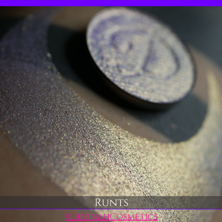 A round eyeshadow pan labeled 'RUNTS' made by Devinah Cosmetics, placed over a large crescent moon swatch of the same shimmering shifty eyeshadow on skin. The eyeshadow has a duochrome multichrome shift between colors purple pink and yellow