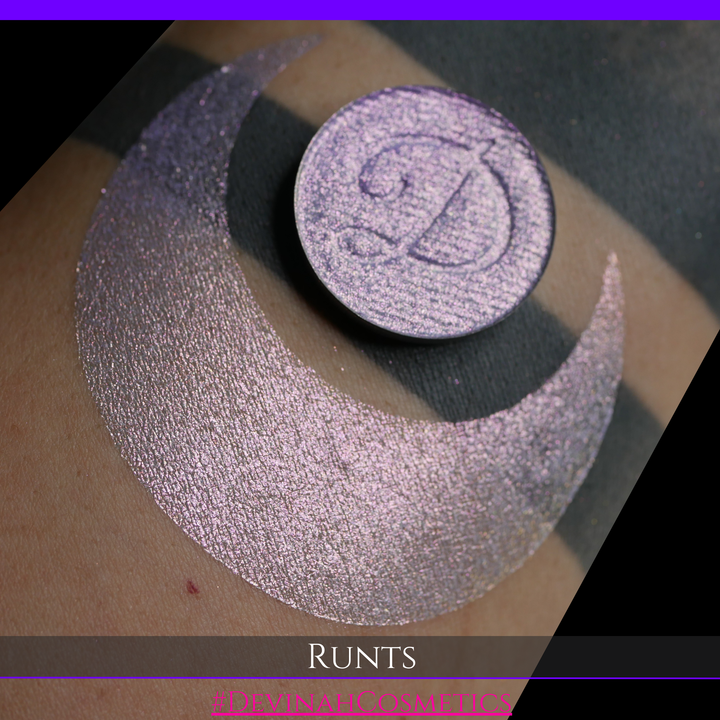 A round eyeshadow pan labeled 'RUNTS' made by Devinah Cosmetics, placed over a large crescent moon swatch of the same shimmering shifty eyeshadow on skin. The eyeshadow has a duochrome multichrome shift between colors purple pink and yellow