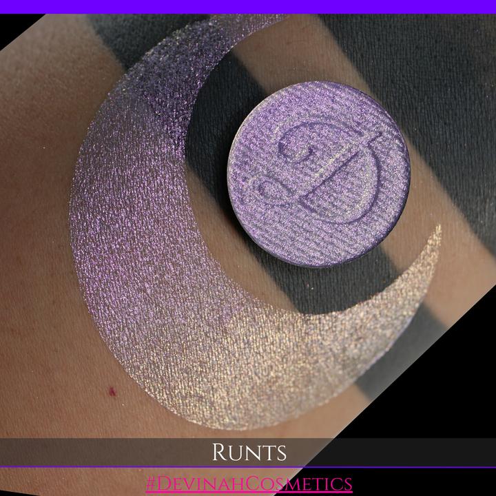 A round eyeshadow pan labeled 'RUNTS' made by Devinah Cosmetics, placed over a large crescent moon swatch of the same shimmering shifty eyeshadow on skin. The eyeshadow has a duochrome multichrome shift between colors purple pink and yellow