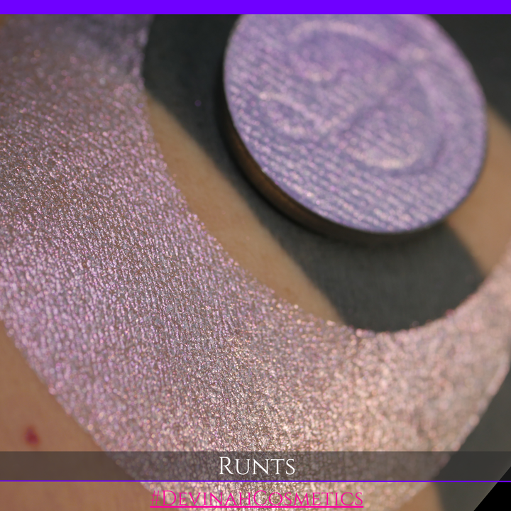 A round eyeshadow pan labeled 'RUNTS' made by Devinah Cosmetics, placed over a large crescent moon swatch of the same shimmering shifty eyeshadow on skin. The eyeshadow has a duochrome multichrome shift between colors purple pink and yellow