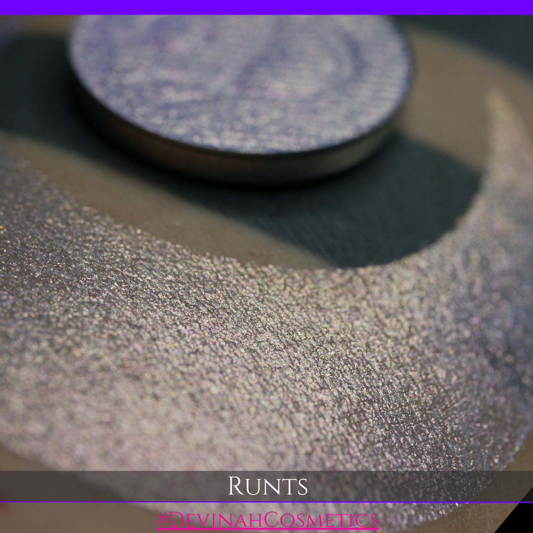 A round eyeshadow pan labeled 'RUNTS' made by Devinah Cosmetics, placed over a large crescent moon swatch of the same shimmering shifty eyeshadow on skin. The eyeshadow has a duochrome multichrome shift between colors purple pink and yellow