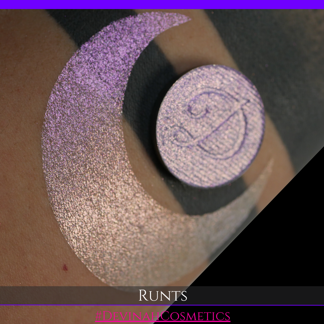 A round eyeshadow pan labeled 'RUNTS' made by Devinah Cosmetics, placed over a large crescent moon swatch of the same shimmering shifty eyeshadow on skin. The eyeshadow has a duochrome multichrome shift between colors purple pink and yellow