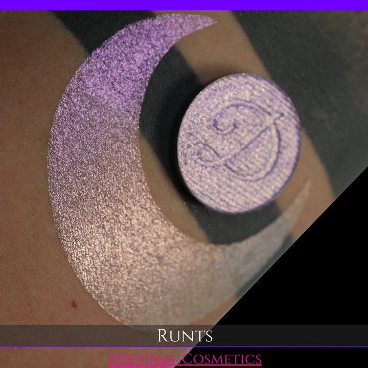 A round eyeshadow pan labeled 'RUNTS' made by Devinah Cosmetics, placed over a large crescent moon swatch of the same shimmering shifty eyeshadow on skin. The eyeshadow has a duochrome multichrome shift between colors purple pink and yellow
