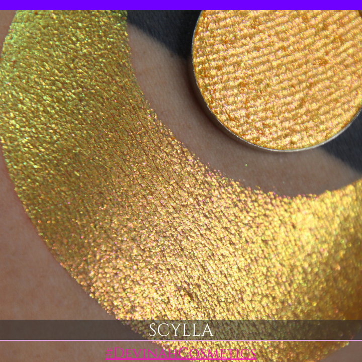 A round eyeshadow pan labeled 'SCYLLA' made by Devinah Cosmetics, placed over a large crescent moon swatch of the same shimmering shifty eyeshadow on skin. The eyeshadow has a duochrome multichrome shift between colors lime green, teal, yellow, gold, orange, pink, red.