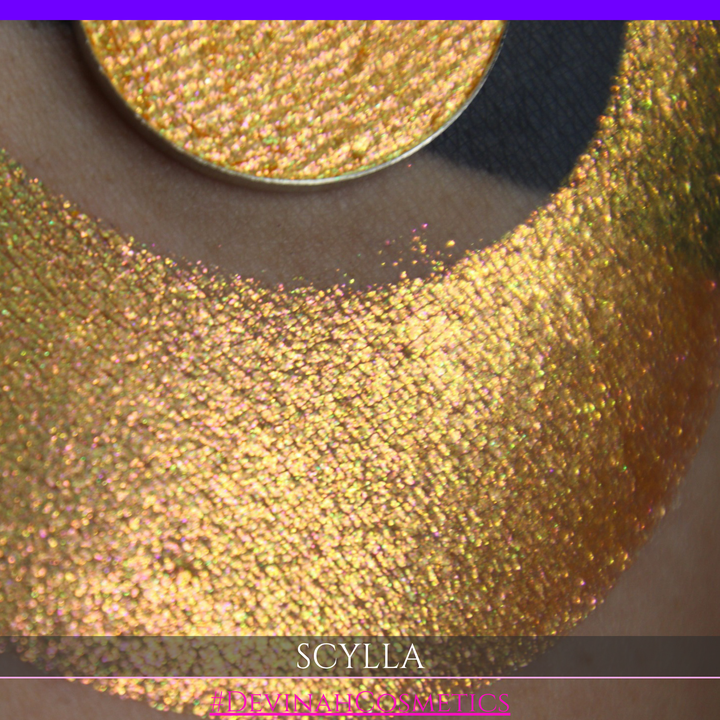 A round eyeshadow pan labeled 'SCYLLA' made by Devinah Cosmetics, placed over a large crescent moon swatch of the same shimmering shifty eyeshadow on skin. The eyeshadow has a duochrome multichrome shift between colors lime green, teal, yellow, gold, orange, pink, red.