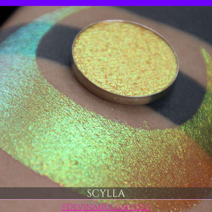 A round eyeshadow pan labeled 'SCYLLA' made by Devinah Cosmetics, placed over a large crescent moon swatch of the same shimmering shifty eyeshadow on skin. The eyeshadow has a duochrome multichrome shift between colors lime green, teal, yellow, gold, orange, pink, red.