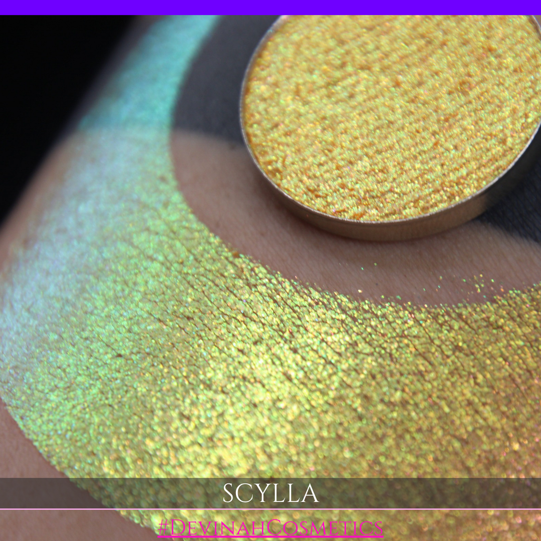 A round eyeshadow pan labeled 'SCYLLA' made by Devinah Cosmetics, placed over a large crescent moon swatch of the same shimmering shifty eyeshadow on skin. The eyeshadow has a duochrome multichrome shift between colors lime green, teal, yellow, gold, orange, pink, red.