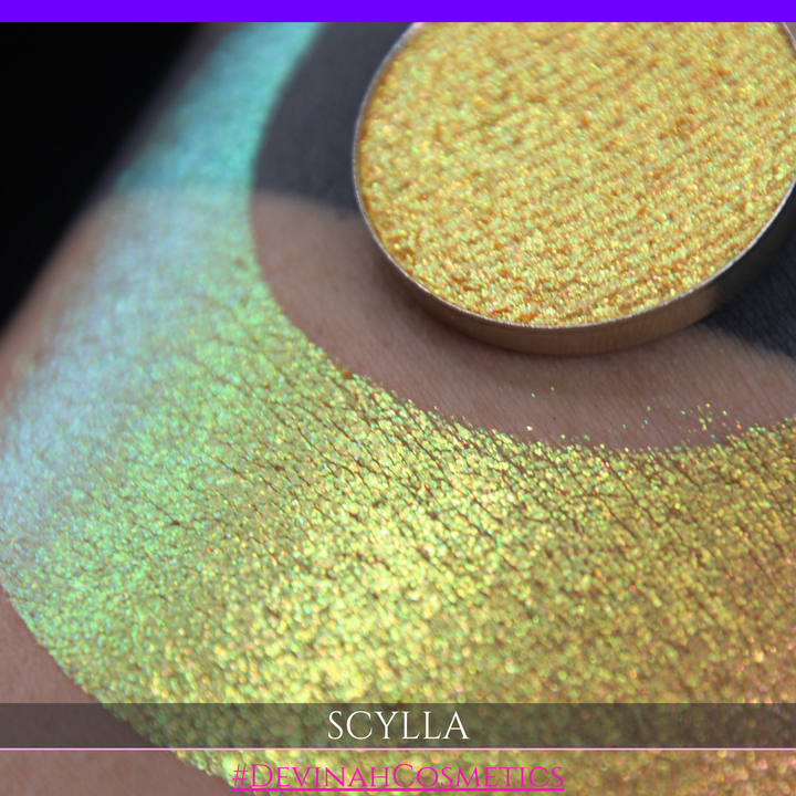 A round eyeshadow pan labeled 'SCYLLA' made by Devinah Cosmetics, placed over a large crescent moon swatch of the same shimmering shifty eyeshadow on skin. The eyeshadow has a duochrome multichrome shift between colors lime green, teal, yellow, gold, orange, pink, red.