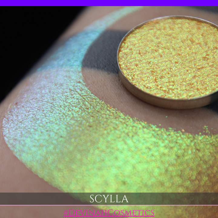 A round eyeshadow pan labeled 'SCYLLA' made by Devinah Cosmetics, placed over a large crescent moon swatch of the same shimmering shifty eyeshadow on skin. The eyeshadow has a duochrome multichrome shift between colors lime green, teal, yellow, gold, orange, pink, red.