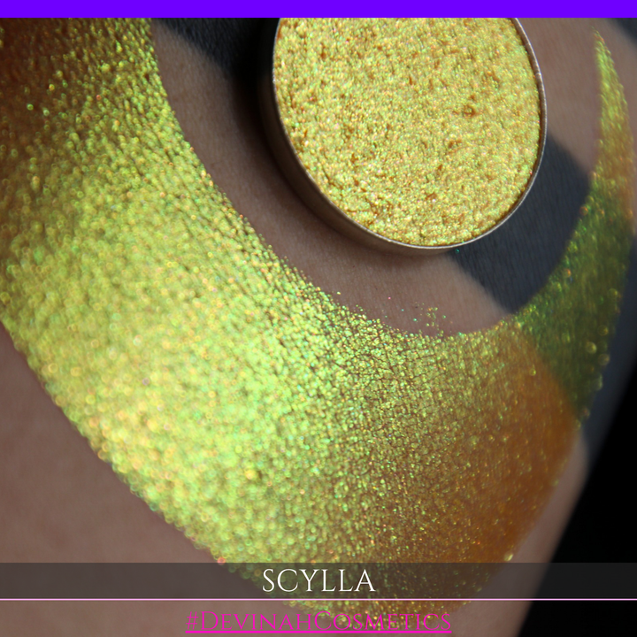 A round eyeshadow pan labeled 'SCYLLA' made by Devinah Cosmetics, placed over a large crescent moon swatch of the same shimmering shifty eyeshadow on skin. The eyeshadow has a duochrome multichrome shift between colors lime green, teal, yellow, gold, orange, pink, red.