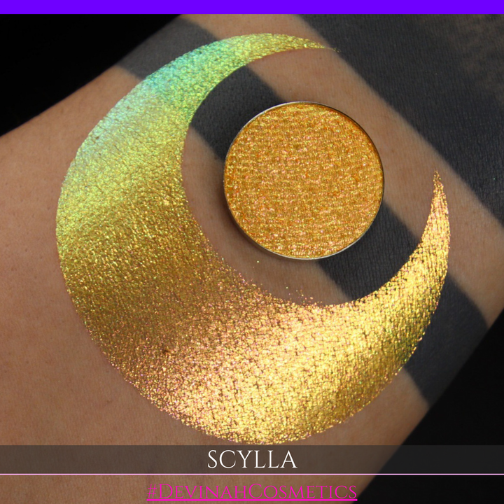 A round eyeshadow pan labeled 'SCYLLA' made by Devinah Cosmetics, placed over a large crescent moon swatch of the same shimmering shifty eyeshadow on skin. The eyeshadow has a duochrome multichrome shift between colors lime green, teal, yellow, gold, orange, pink, red.