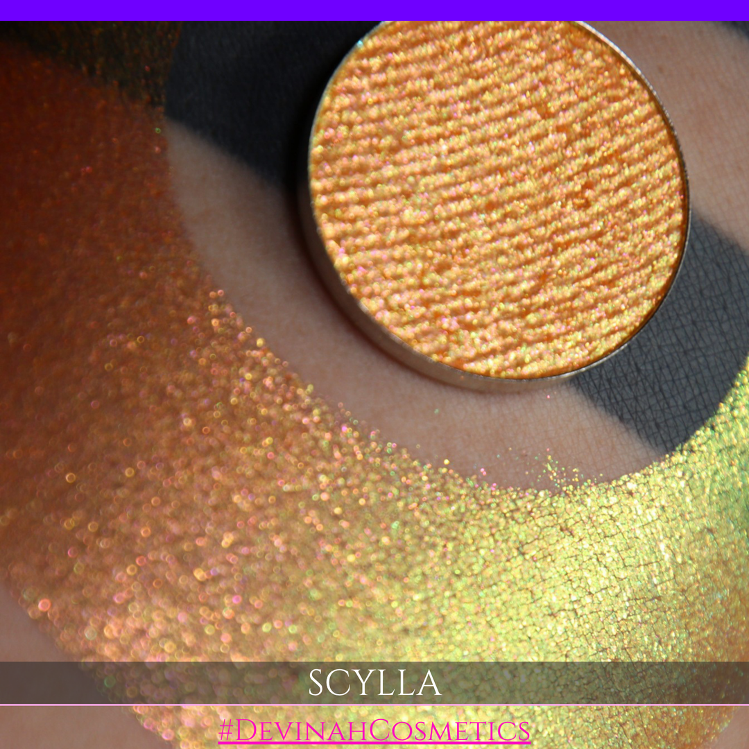 A round eyeshadow pan labeled 'SCYLLA' made by Devinah Cosmetics, placed over a large crescent moon swatch of the same shimmering shifty eyeshadow on skin. The eyeshadow has a duochrome multichrome shift between colors lime green, teal, yellow, gold, orange, pink, red.