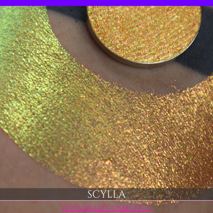A round eyeshadow pan labeled 'SCYLLA' made by Devinah Cosmetics, placed over a large crescent moon swatch of the same shimmering shifty eyeshadow on skin. The eyeshadow has a duochrome multichrome shift between colors lime green, teal, yellow, gold, orange, pink, red.