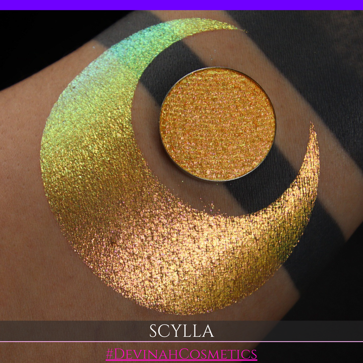 A round eyeshadow pan labeled 'SCYLLA' made by Devinah Cosmetics, placed over a large crescent moon swatch of the same shimmering shifty eyeshadow on skin. The eyeshadow has a duochrome multichrome shift between colors lime green, teal, yellow, gold, orange, pink, red.