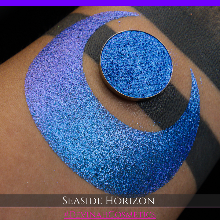 A round eyeshadow pan labeled 'SEASIDE HORIZON' made by Devinah Cosmetics, placed over a large crescent moon swatch of the same shimmering shifty eyeshadow on skin. The eyeshadow has a shift between colors purple, blue.