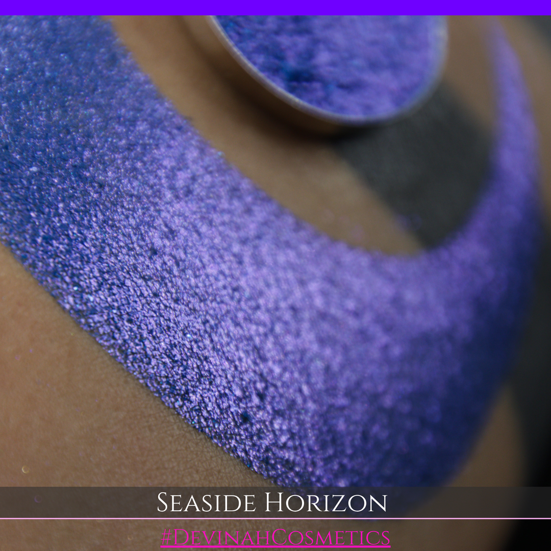 A round eyeshadow pan labeled 'SEASIDE HORIZON' made by Devinah Cosmetics, placed over a large crescent moon swatch of the same shimmering shifty eyeshadow on skin. The eyeshadow has a shift between colors purple, blue.