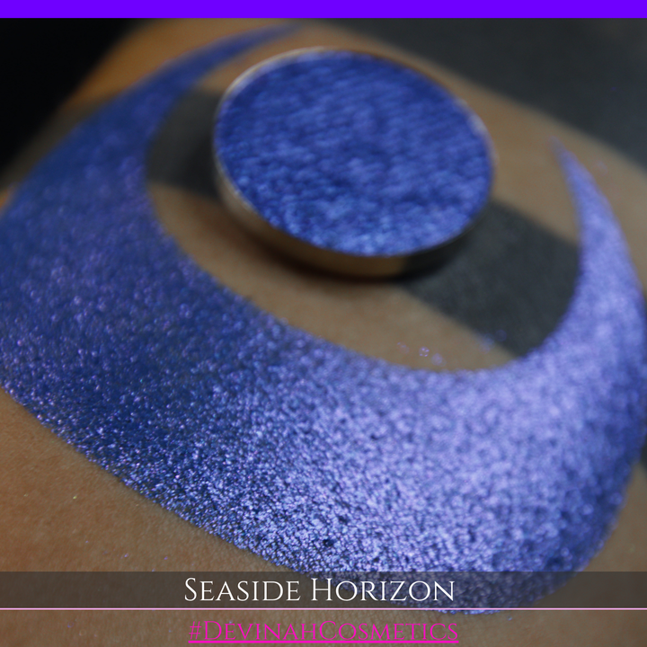 A round eyeshadow pan labeled 'SEASIDE HORIZON' made by Devinah Cosmetics, placed over a large crescent moon swatch of the same shimmering shifty eyeshadow on skin. The eyeshadow has a shift between colors purple, blue.
