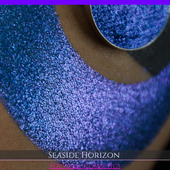 A round eyeshadow pan labeled 'SEASIDE HORIZON' made by Devinah Cosmetics, placed over a large crescent moon swatch of the same shimmering shifty eyeshadow on skin. The eyeshadow has a shift between colors purple, blue.