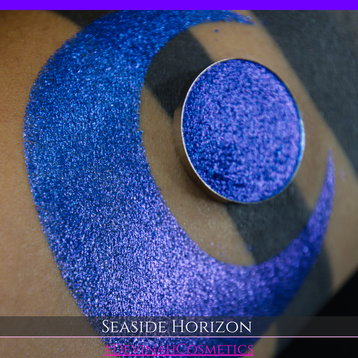 A round eyeshadow pan labeled 'SEASIDE HORIZON' made by Devinah Cosmetics, placed over a large crescent moon swatch of the same shimmering shifty eyeshadow on skin. The eyeshadow has a shift between colors purple, blue.