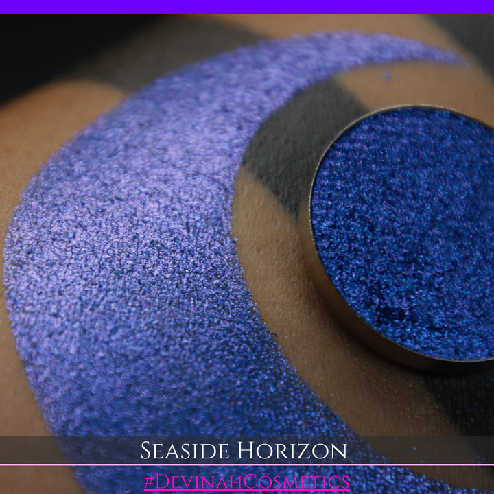A round eyeshadow pan labeled 'SEASIDE HORIZON' made by Devinah Cosmetics, placed over a large crescent moon swatch of the same shimmering shifty eyeshadow on skin. The eyeshadow has a shift between colors purple, blue.