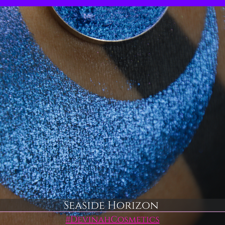 A round eyeshadow pan labeled 'SEASIDE HORIZON' made by Devinah Cosmetics, placed over a large crescent moon swatch of the same shimmering shifty eyeshadow on skin. The eyeshadow has a shift between colors purple, blue.