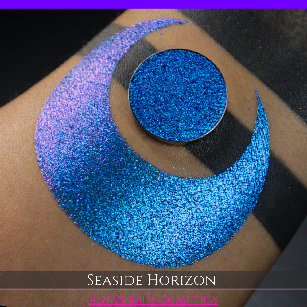 A round eyeshadow pan labeled 'SEASIDE HORIZON' made by Devinah Cosmetics, placed over a large crescent moon swatch of the same shimmering shifty eyeshadow on skin. The eyeshadow has a shift between colors purple, blue.