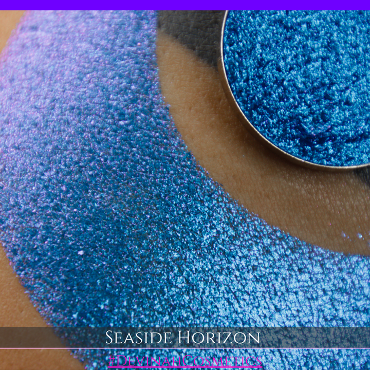 A round eyeshadow pan labeled 'SEASIDE HORIZON' made by Devinah Cosmetics, placed over a large crescent moon swatch of the same shimmering shifty eyeshadow on skin. The eyeshadow has a shift between colors purple, blue.