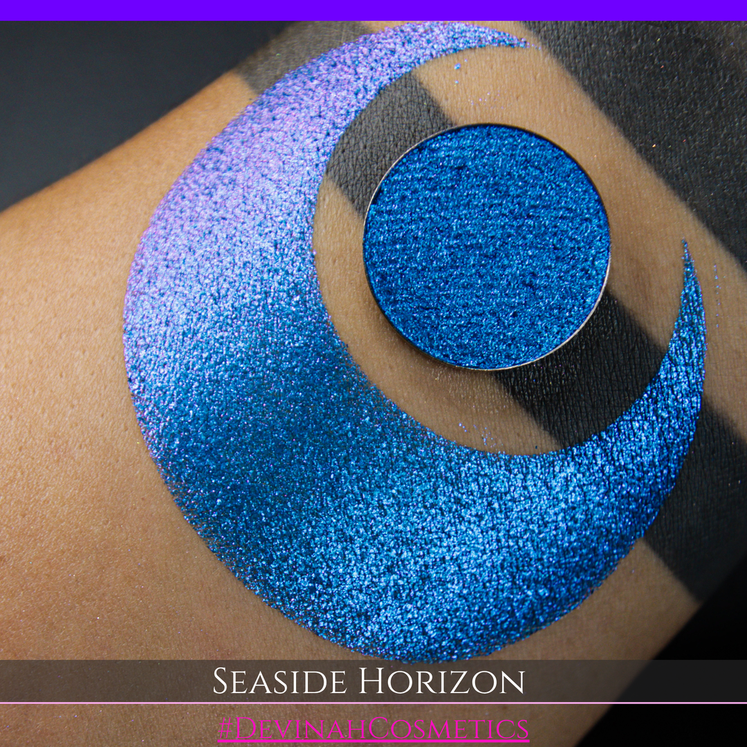 A round eyeshadow pan labeled 'SEASIDE HORIZON' made by Devinah Cosmetics, placed over a large crescent moon swatch of the same shimmering shifty eyeshadow on skin. The eyeshadow has a shift between colors purple, blue.