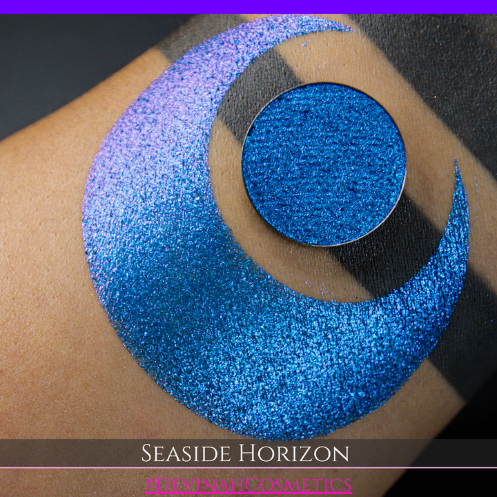 A round eyeshadow pan labeled 'SEASIDE HORIZON' made by Devinah Cosmetics, placed over a large crescent moon swatch of the same shimmering shifty eyeshadow on skin. The eyeshadow has a shift between colors purple, blue.