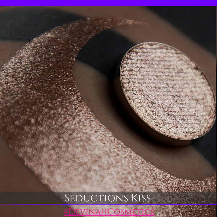 A round eyeshadow pan labeled 'SEDUCTIONS KISS' made by Devinah Cosmetics, placed over a large crescent moon swatch of the same shimmering shifty eyeshadow on skin. The eyeshadow has colors brown, tan, creme sparkle shimmer.