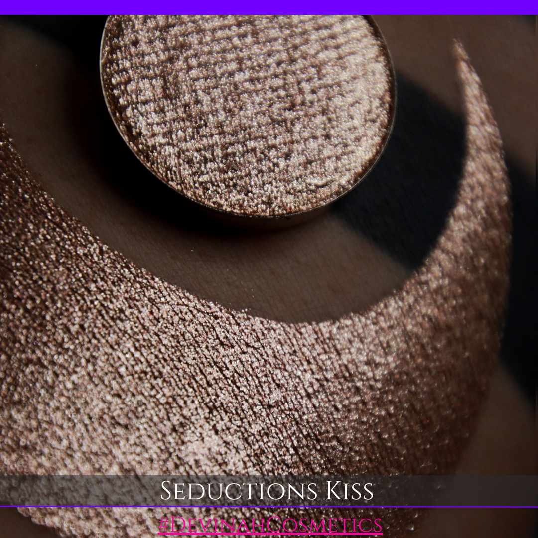 A round eyeshadow pan labeled 'SEDUCTIONS KISS' made by Devinah Cosmetics, placed over a large crescent moon swatch of the same shimmering shifty eyeshadow on skin. The eyeshadow has colors brown, tan, creme sparkle shimmer.