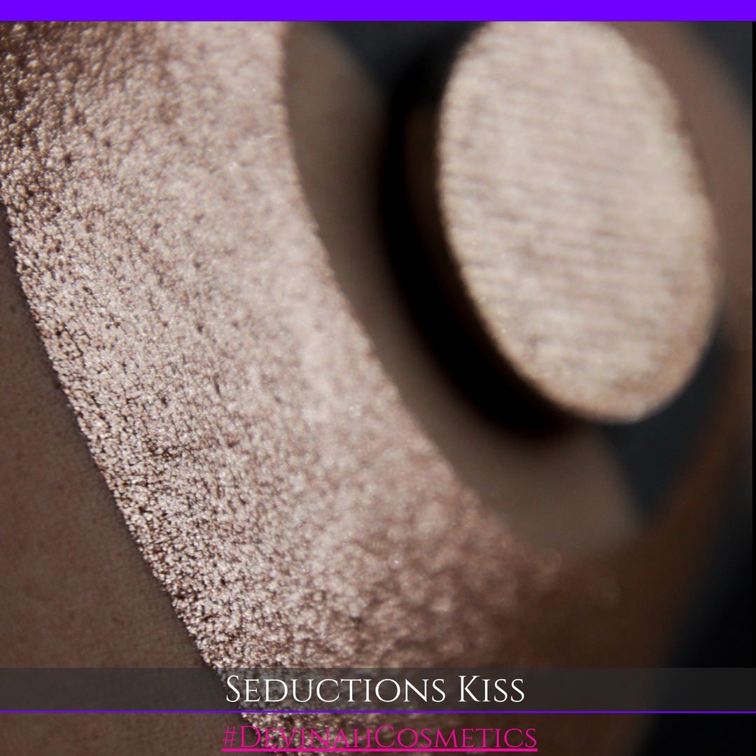 A round eyeshadow pan labeled 'SEDUCTIONS KISS' made by Devinah Cosmetics, placed over a large crescent moon swatch of the same shimmering shifty eyeshadow on skin. The eyeshadow has colors brown, tan, creme sparkle shimmer.