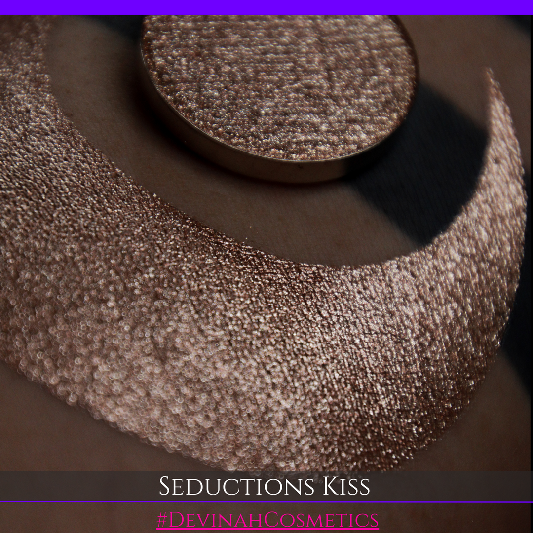A round eyeshadow pan labeled 'SEDUCTIONS KISS' made by Devinah Cosmetics, placed over a large crescent moon swatch of the same shimmering shifty eyeshadow on skin. The eyeshadow has colors brown, tan, creme sparkle shimmer.