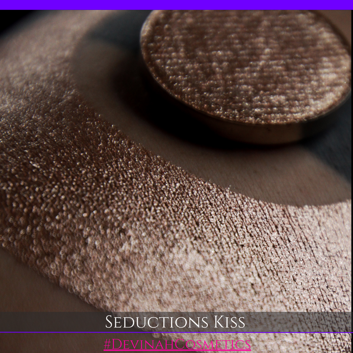 A round eyeshadow pan labeled 'SEDUCTIONS KISS' made by Devinah Cosmetics, placed over a large crescent moon swatch of the same shimmering shifty eyeshadow on skin. The eyeshadow has colors brown, tan, creme sparkle shimmer.