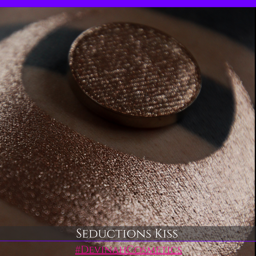 A round eyeshadow pan labeled 'SEDUCTIONS KISS' made by Devinah Cosmetics, placed over a large crescent moon swatch of the same shimmering shifty eyeshadow on skin. The eyeshadow has colors brown, tan, creme sparkle shimmer.