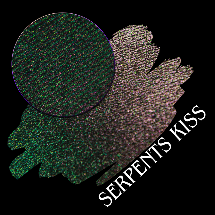 A round eyeshadow pan labeled 'SERPENTS KISS' made by Devinah Cosmetics, placed over a large crescent moon swatch of the same shimmering shifty eyeshadow on skin. The eyeshadow has a duochrome multichrome shift between colors greeen, olive brown, red.