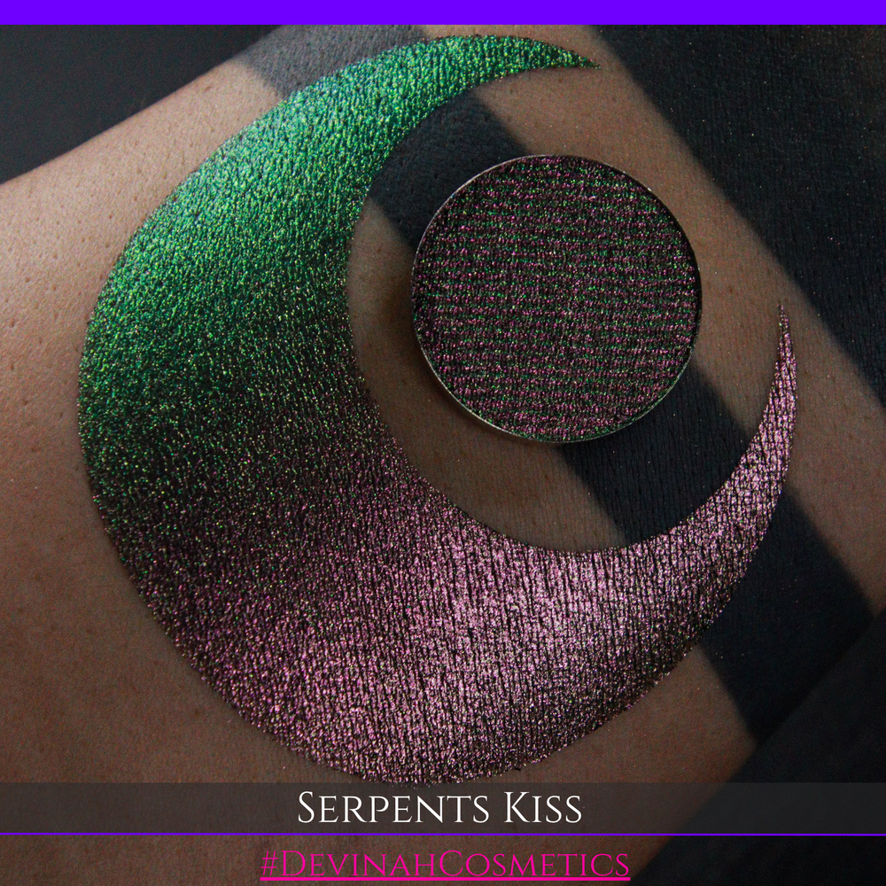 A round eyeshadow pan labeled 'SERPENTS KISS' made by Devinah Cosmetics, placed over a large crescent moon swatch of the same shimmering shifty eyeshadow on skin. The eyeshadow has a duochrome multichrome shift between colors greeen, olive brown, red.
