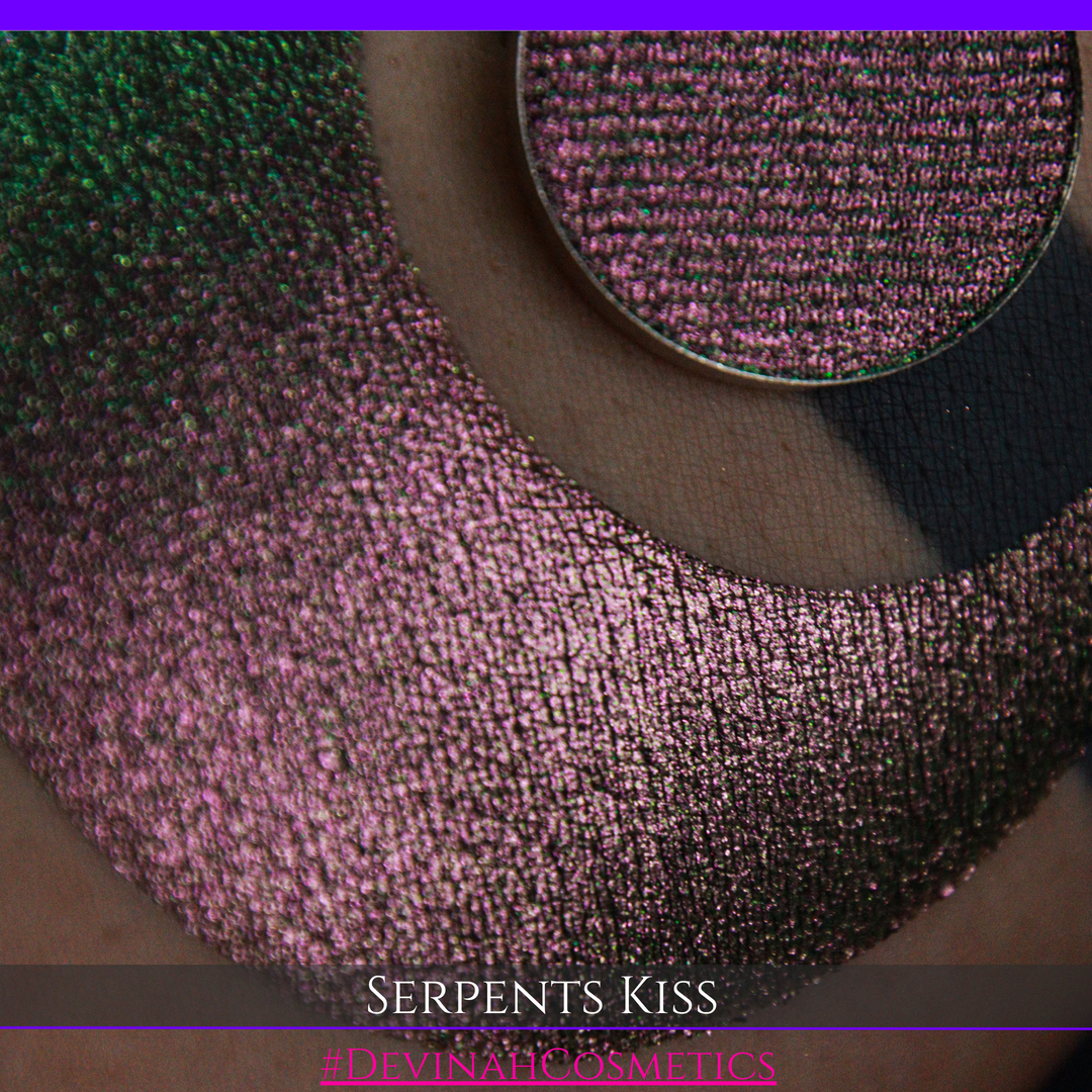 A round eyeshadow pan labeled 'SERPENTS KISS' made by Devinah Cosmetics, placed over a large crescent moon swatch of the same shimmering shifty eyeshadow on skin. The eyeshadow has a duochrome multichrome shift between colors greeen, olive brown, red.
