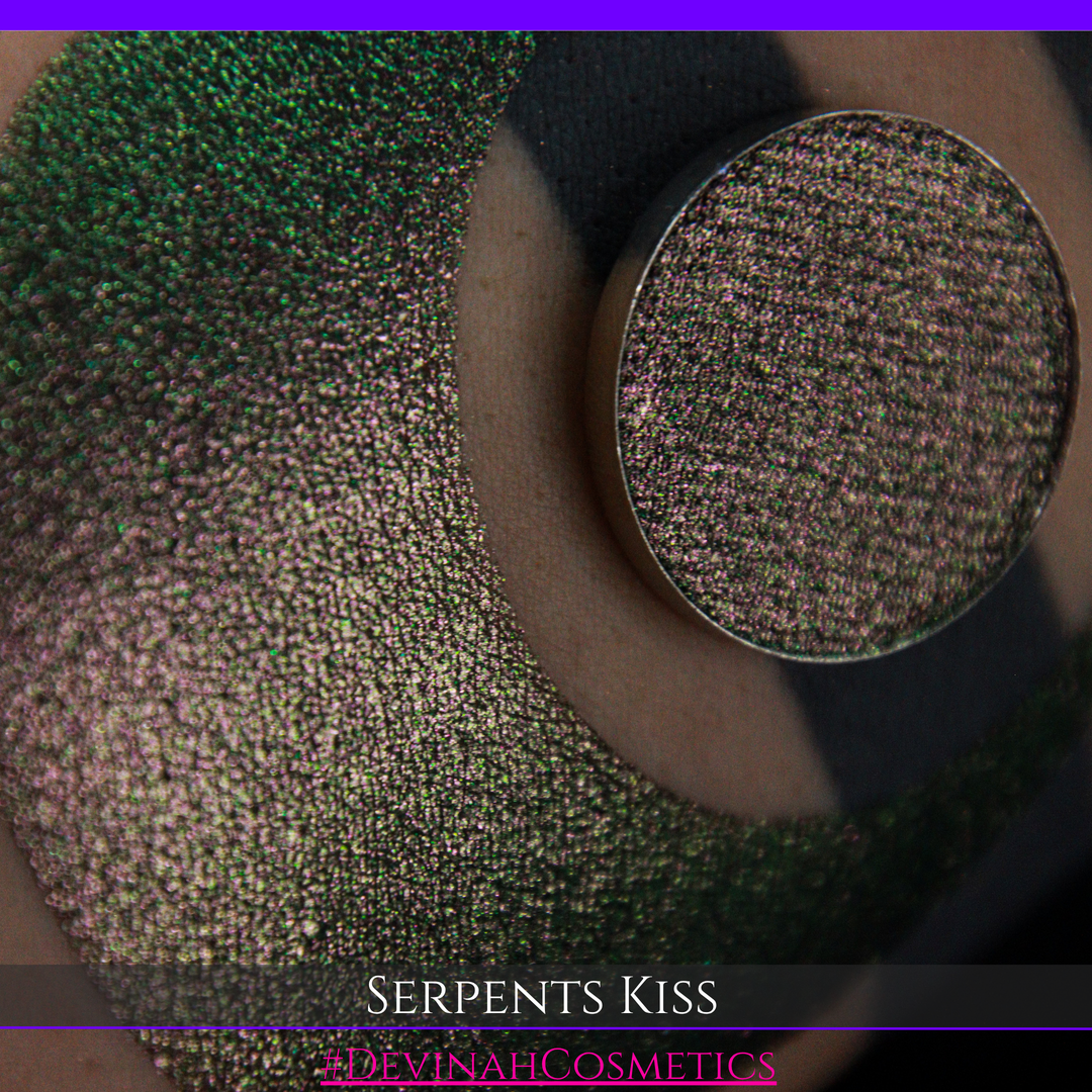 A round eyeshadow pan labeled 'SERPENTS KISS' made by Devinah Cosmetics, placed over a large crescent moon swatch of the same shimmering shifty eyeshadow on skin. The eyeshadow has a duochrome multichrome shift between colors greeen, olive brown, red.