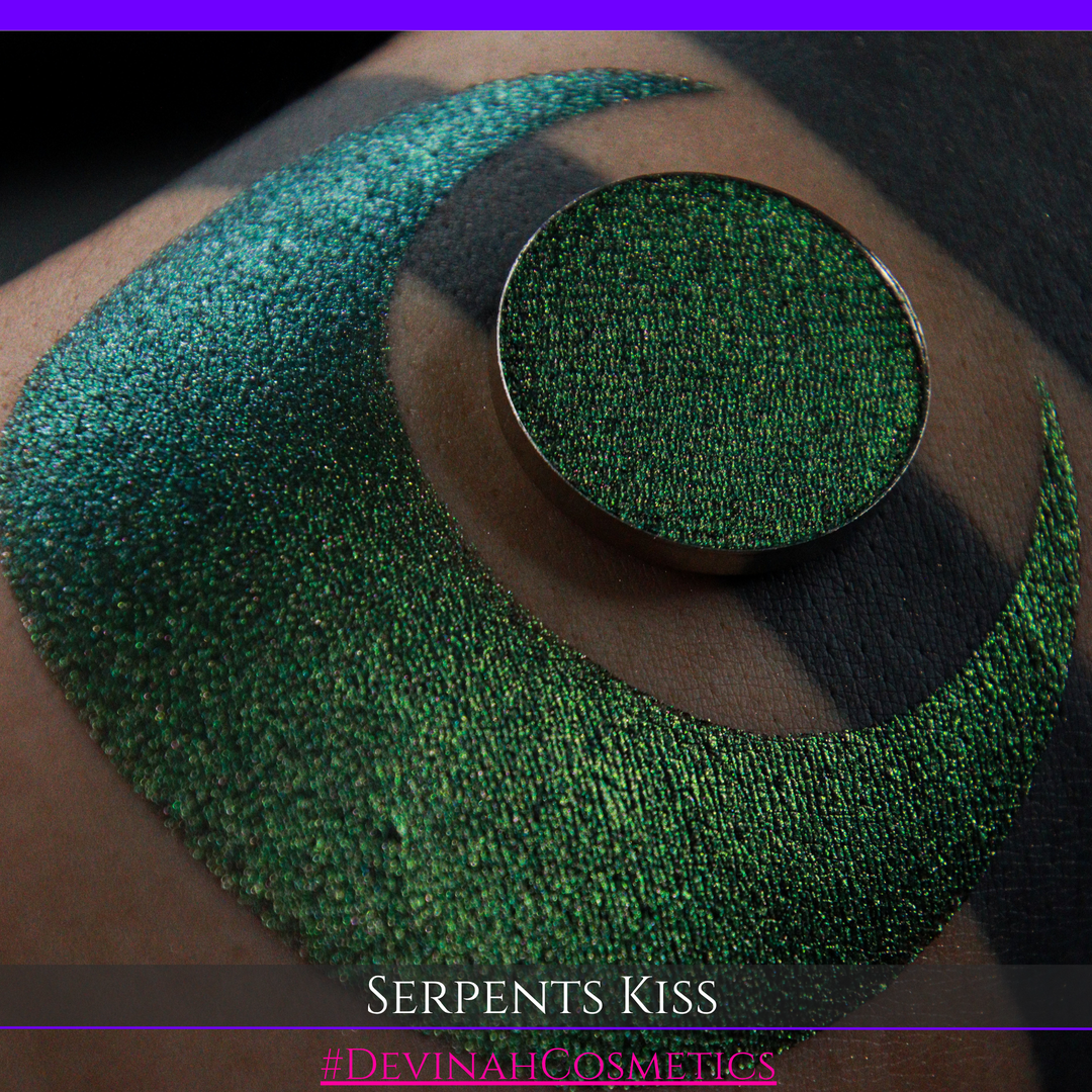 A round eyeshadow pan labeled 'SERPENTS KISS' made by Devinah Cosmetics, placed over a large crescent moon swatch of the same shimmering shifty eyeshadow on skin. The eyeshadow has a duochrome multichrome shift between colors greeen, olive brown, red.