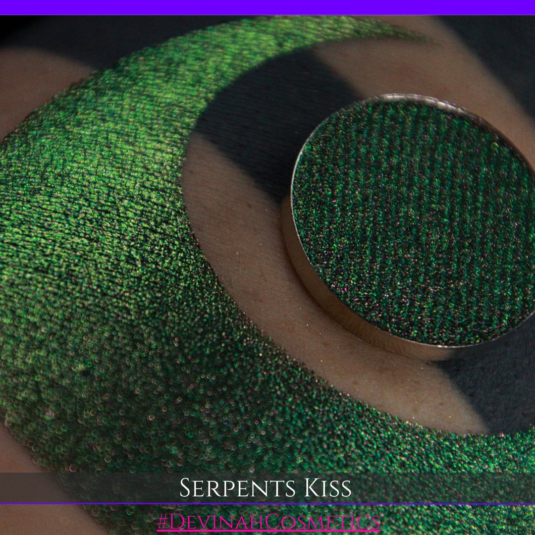 A round eyeshadow pan labeled 'SERPENTS KISS' made by Devinah Cosmetics, placed over a large crescent moon swatch of the same shimmering shifty eyeshadow on skin. The eyeshadow has a duochrome multichrome shift between colors greeen, olive brown, red.