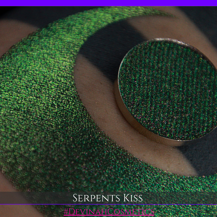 A round eyeshadow pan labeled 'SERPENTS KISS' made by Devinah Cosmetics, placed over a large crescent moon swatch of the same shimmering shifty eyeshadow on skin. The eyeshadow has a duochrome multichrome shift between colors greeen, olive brown, red.