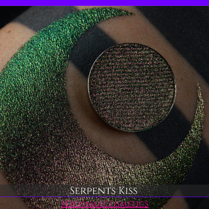 A round eyeshadow pan labeled 'SERPENTS KISS' made by Devinah Cosmetics, placed over a large crescent moon swatch of the same shimmering shifty eyeshadow on skin. The eyeshadow has a duochrome multichrome shift between colors greeen, olive brown, red.